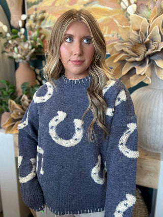A woman wearing a navy blue knit sweater with a white horseshoe pattern, standing in front of a decorative floral background.