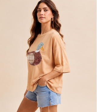 Woman wearing a camel Mallard Duck Patchwork Knit Top with plaid and floral duck appliqué and denim shorts.