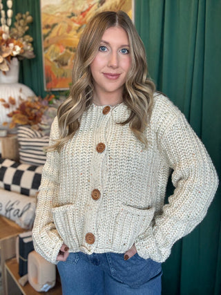 Woman wearing an oatmeal chunky knit cardigan with multicolor confetti flecks, oversized wood-look buttons, and front patch pockets.