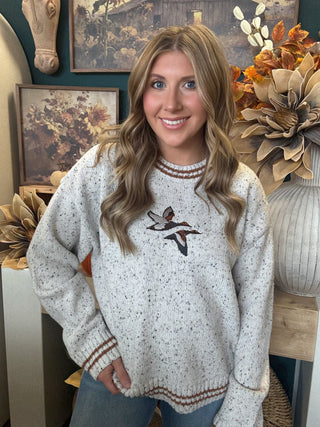 Woman wearing a speckled oatmeal sweater with embroidered flying mallards and rust-brown striped trim
