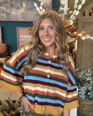 Woman wearing a mustard, cream, rust, forest green, light blue, and burgundy striped button-front sweater.