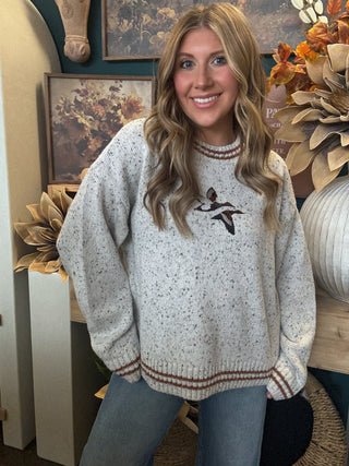 Woman wearing a speckled oatmeal sweater with embroidered flying mallards and rust-brown striped trim