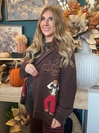 Woman wearing a chocolate-brown open-front cardigan with embroidered “Howdy” script and colorful cowboy motifs