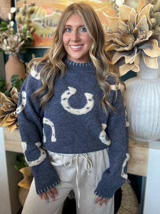 A woman wearing a navy blue knit sweater with cream horseshoe patterns, paired with light beige sweatpants, standing in a decorated indoor setting with large dried flowers.