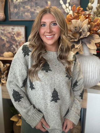 Woman wearing a heathered oatmeal-gray sweater with raised dark evergreen-tree motifs