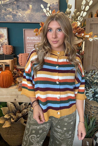 Woman wearing a mustard, cream, rust, forest green, light blue, and burgundy striped button-front sweater.