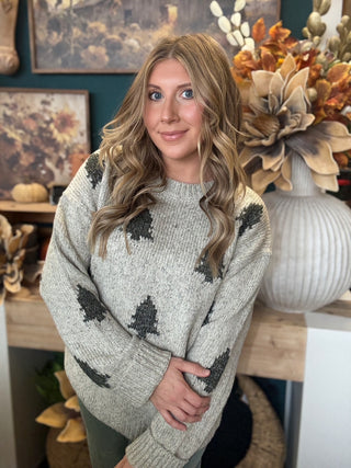 Woman wearing a heathered oatmeal-gray sweater with raised dark evergreen-tree motifs