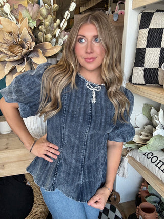 Woman wearing a washed-charcoal peplum top with puff sleeves and pearl bow neckline detail