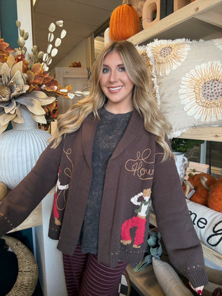 Woman wearing a chocolate-brown open-front cardigan with embroidered “Howdy” script and colorful cowboy motifs