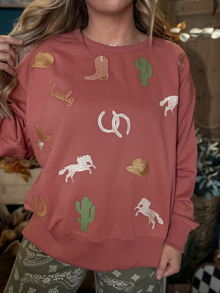 Woman wearing a dusty-rose sweatshirt with embroidered cowboy hats, boots, cacti, horses, and horseshoes