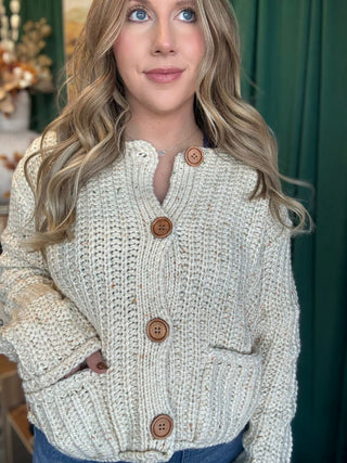 Woman wearing an oatmeal chunky knit cardigan with multicolor confetti flecks, oversized wood-look buttons, and front patch pockets.