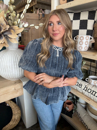 Woman wearing a washed-charcoal peplum top with puff sleeves and pearl bow neckline detail