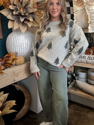 Woman wearing a heathered oatmeal-gray sweater with raised dark evergreen-tree motifs