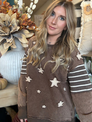 Woman wearing a chocolate vintage-wash pullover with cream appliqué stars and striped sleeves