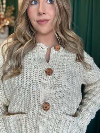 Woman wearing an oatmeal chunky knit cardigan with multicolor confetti flecks, oversized wood-look buttons, and front patch pockets.