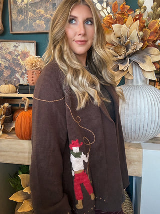 Woman wearing a chocolate-brown open-front cardigan with embroidered “Howdy” script and colorful cowboy motifs