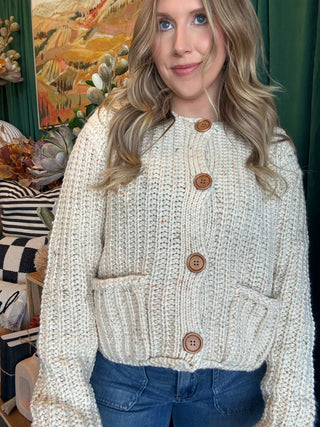 Woman wearing an oatmeal chunky knit cardigan with multicolor confetti flecks, oversized wood-look buttons, and front patch pockets.