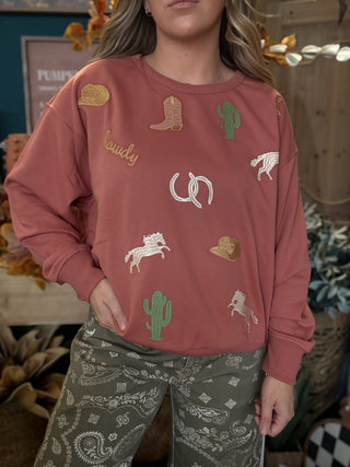 Woman wearing a dusty-rose sweatshirt with embroidered cowboy hats, boots, cacti, horses, and horseshoes