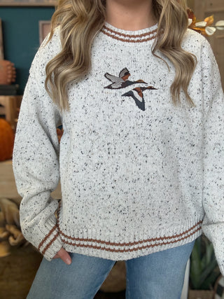 Woman wearing a speckled oatmeal sweater with embroidered flying mallards and rust-brown striped trim