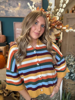 Woman wearing a mustard, cream, rust, forest green, light blue, and burgundy striped button-front sweater.