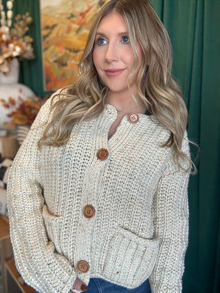 Woman wearing an oatmeal chunky knit cardigan with multicolor confetti flecks, oversized wood-look buttons, and front patch pockets.