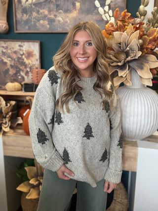 Woman wearing a heathered oatmeal-gray sweater with raised dark evergreen-tree motifs