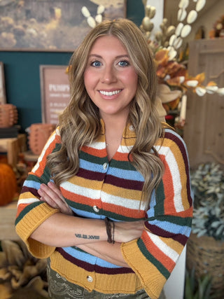 Woman wearing a mustard, cream, rust, forest green, light blue, and burgundy striped button-front sweater.