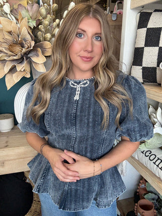 Woman wearing a washed-charcoal peplum top with puff sleeves and pearl bow neckline detail