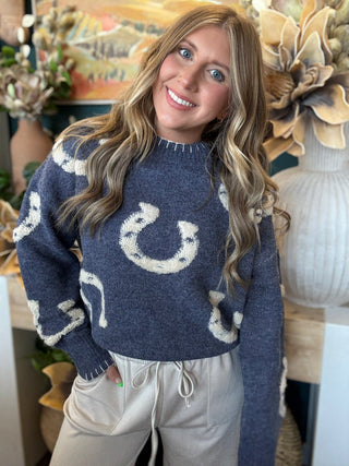 A woman smiling while wearing a navy blue knit sweater with large white horseshoe patterns, paired with light beige drawstring sweatpants in a decorated indoor setting.