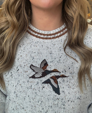 Woman wearing a speckled oatmeal sweater with embroidered flying mallards and rust-brown striped trim