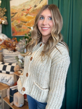 Woman wearing an oatmeal chunky knit cardigan with multicolor confetti flecks, oversized wood-look buttons, and front patch pockets.