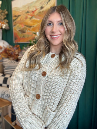 Woman wearing an oatmeal chunky knit cardigan with multicolor confetti flecks, oversized wood-look buttons, and front patch pockets.