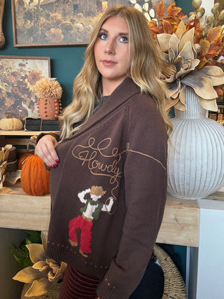 Woman wearing a chocolate-brown open-front cardigan with embroidered “Howdy” script and colorful cowboy motifs
