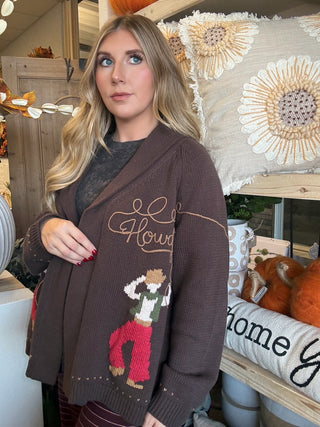 Woman wearing a chocolate-brown open-front cardigan with embroidered “Howdy” script and colorful cowboy motifs