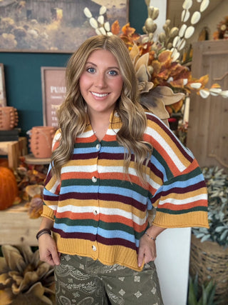 Woman wearing a mustard, cream, rust, forest green, light blue, and burgundy striped button-front sweater.