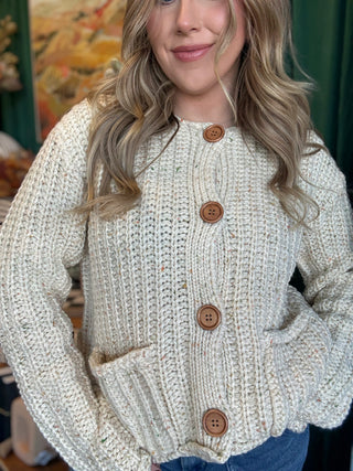 Woman wearing an oatmeal chunky knit cardigan with multicolor confetti flecks, oversized wood-look buttons, and front patch pockets.