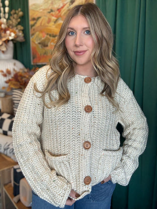 Woman wearing an oatmeal chunky knit cardigan with multicolor confetti flecks, oversized wood-look buttons, and front patch pockets.