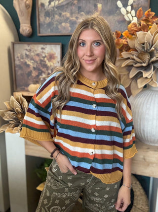 Woman wearing a mustard, cream, rust, forest green, light blue, and burgundy striped button-front sweater.
