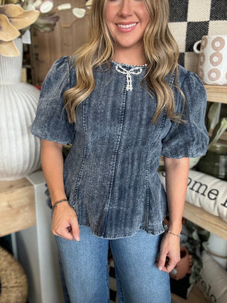 Woman wearing a washed-charcoal peplum top with puff sleeves and pearl bow neckline detail