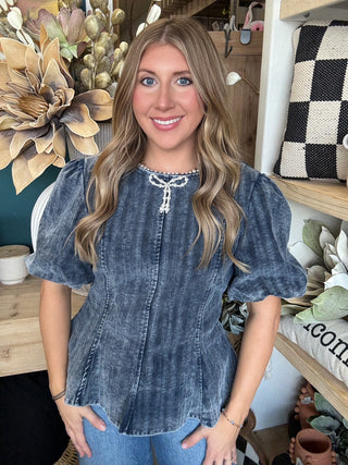 Woman wearing a washed-charcoal peplum top with puff sleeves and pearl bow neckline detail