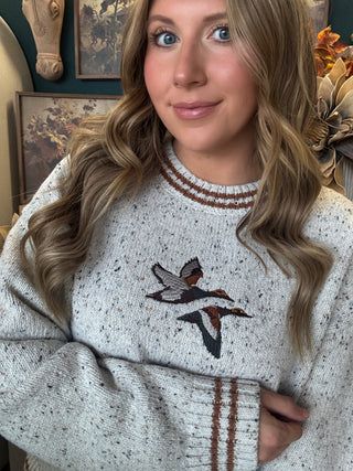 Woman wearing a speckled oatmeal sweater with embroidered flying mallards and rust-brown striped trim