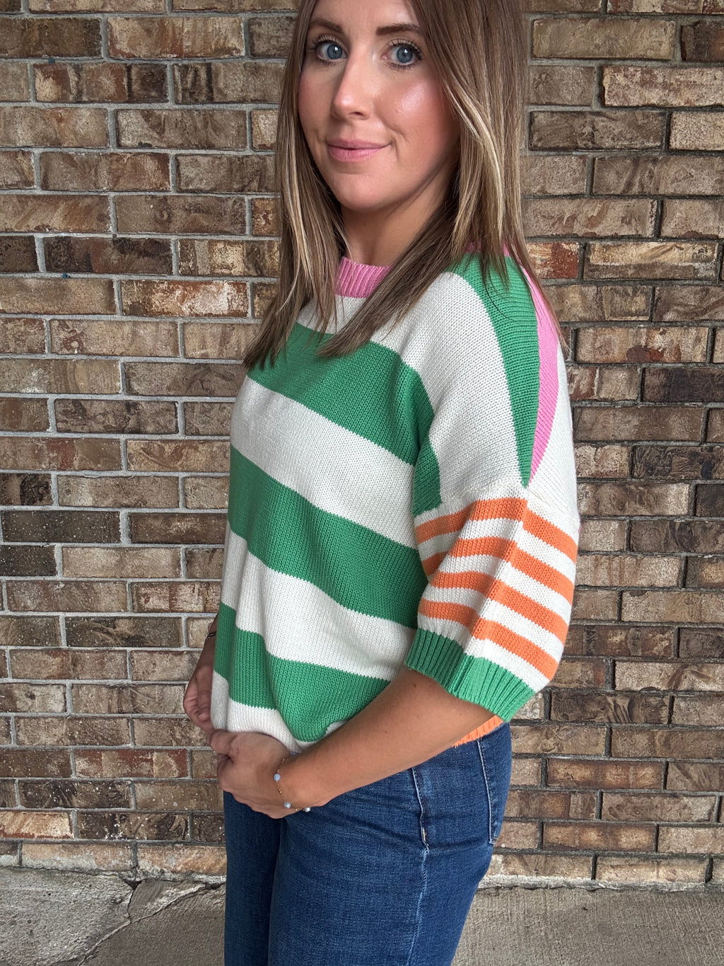 Woman wearing a green and ivory striped lightweight sweater with orange striped sleeves and pink neckline