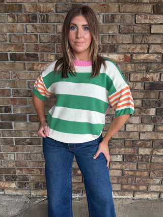 Woman wearing a green and ivory striped lightweight sweater with orange striped sleeves and pink neckline