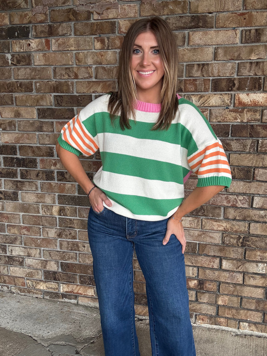 Woman wearing a green and ivory striped lightweight sweater with orange striped sleeves and pink neckline