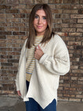 Woman wearing an oversized ivory open knit cardigan layered over a striped sweater and dark wash jeans