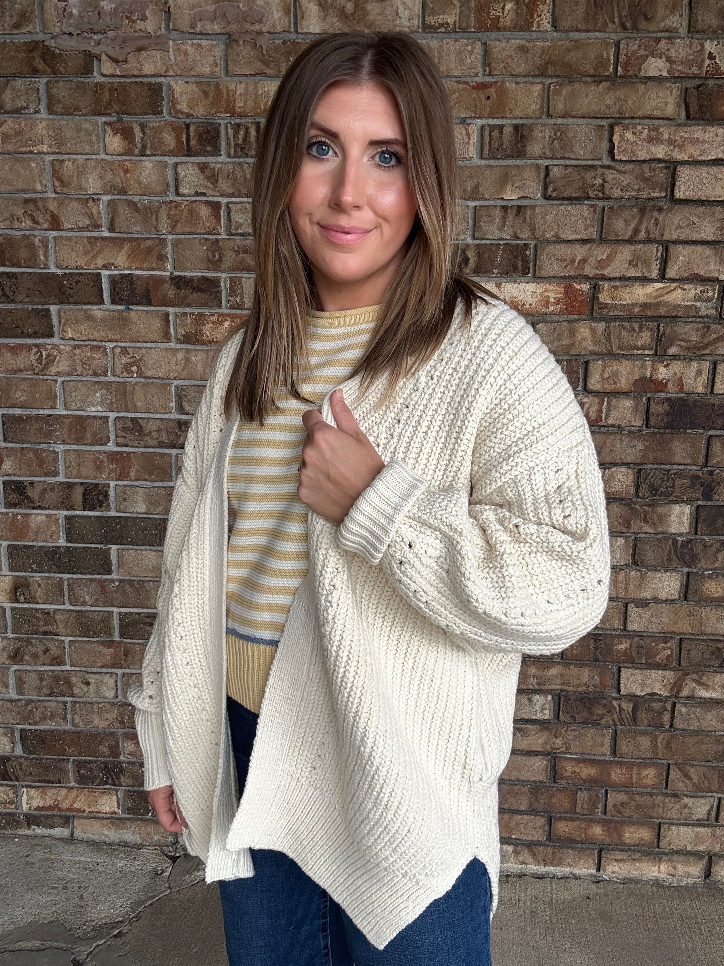 Woman wearing an oversized ivory open knit cardigan layered over a striped sweater and dark wash jeans