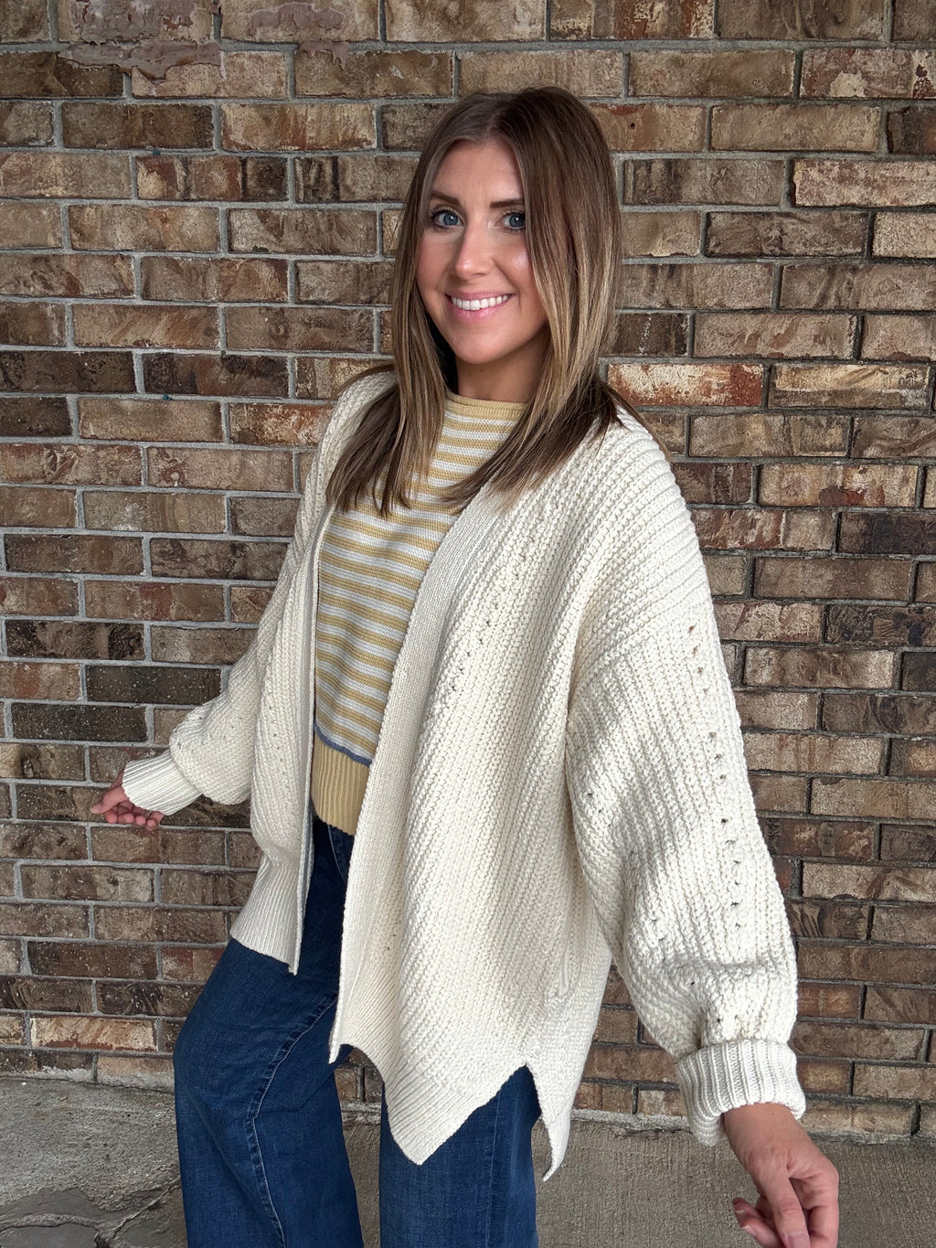 Woman wearing an oversized ivory open knit cardigan layered over a striped sweater and dark wash jeans