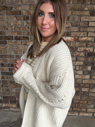 Woman wearing an oversized ivory open knit cardigan layered over a striped sweater and dark wash jeans