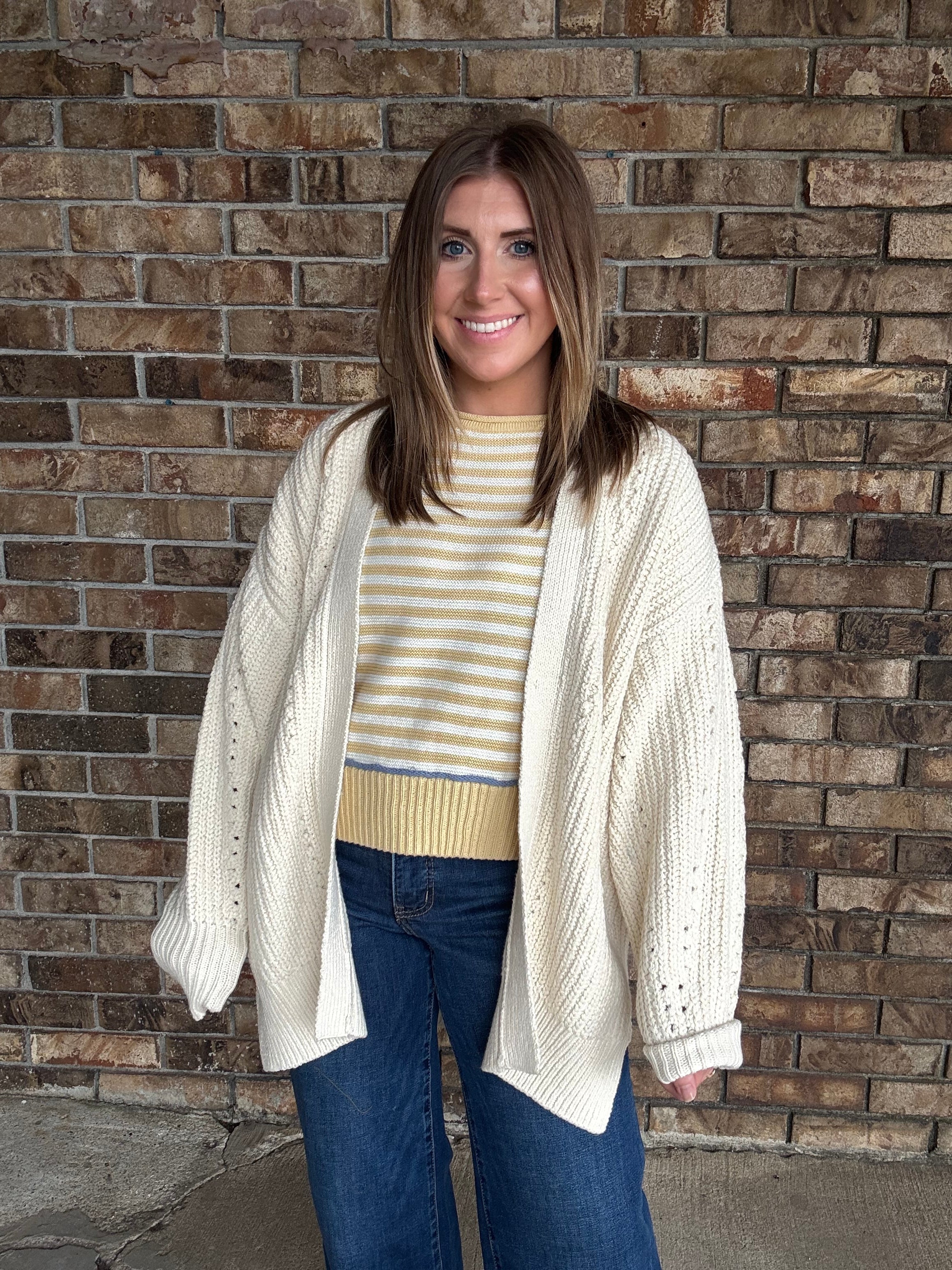 Woman wearing an oversized ivory open knit cardigan layered over a striped sweater and dark wash jeans