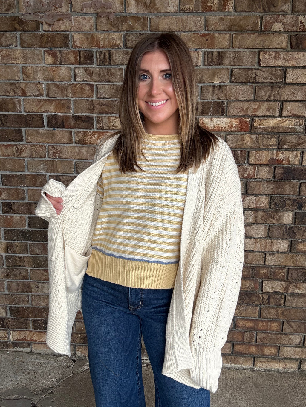 Woman wearing an oversized ivory open knit cardigan layered over a striped sweater and dark wash jeans